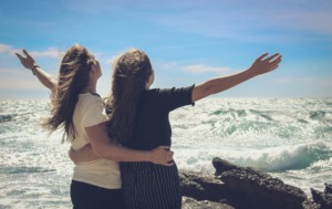 Two women standing at the edge of the ocean with their arms outstretched to the sky, celebrating their freedom, emotional healing, and mutual support on the hero's journey to the authentic self.