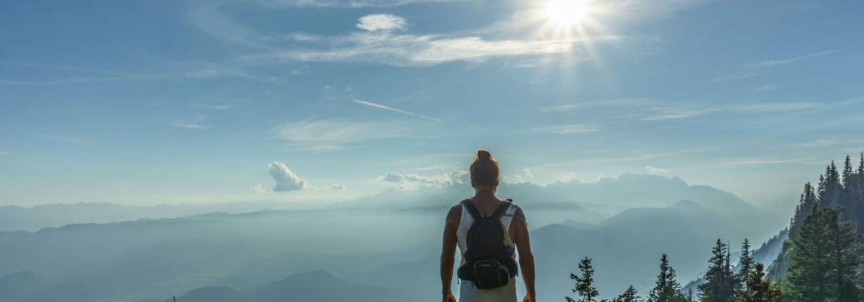 A solitary hiker with a backpack standing on a mountain peak looking out over misty valleys and layered hills under a bright sun, representing the hero's journey to the Authentic Self for highly sensitive introverts.