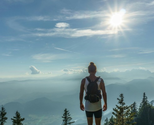 A solitary hiker with a backpack standing on a mountain peak looking out over misty valleys and layered hills under a bright sun, representing the hero's journey to the Authentic Self for highly sensitive introverts.