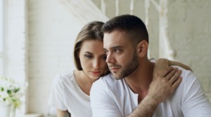 A concerned couple sitting closely, touching hands; the husband looks ahead while the wife rests behind him looking downward, reflecting shared vulnerability and processing relational challenges together.