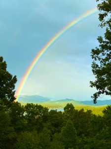 A brilliant, vibrant rainbow arching gracefully across a soft blue sky over lush green forests, distant mountain peaks, and a peaceful lake after a storm.