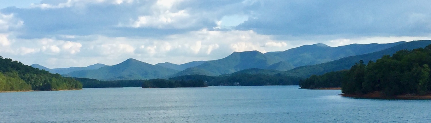 Landscape of Lake Chatuge in the Blue Ridge Mountains of North Georgia where therapist Benita Esposito leads HSP Retreats. The beautiful waters and distant blue mountain peaks reflect the serene setting where clients experience somatic healing. Large white clouds fill the sky above lush green shorelines, capturing the transition from deep emotional isolation and darkness into restoration and inner peace.