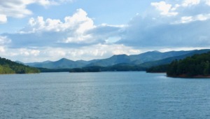 Landscape of Lake Chatuge in the Blue Ridge Mountains of Georgia where therapist Benita Esposito leads HSP Retreats. The beautiful waters and distant blue mountain peaks reflect the serene setting where Kyle experienced somatic healing. Large white clouds fill the sky above lush green shorelines, capturing the transition from deep emotional isolation and darkness into lasting restoration.