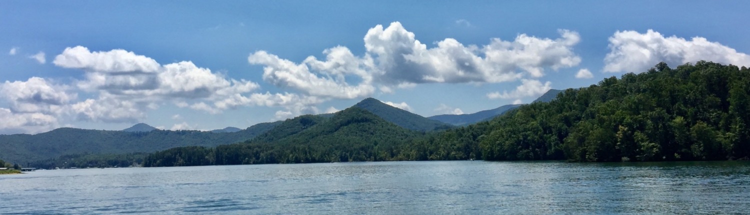 Scenic view of Lake Chatuge in Young Harris, Georgia, surrounded by green forested mountains under a bright blue sky with white clouds where HSP Retreats are hosted by Benita A. Esposito.