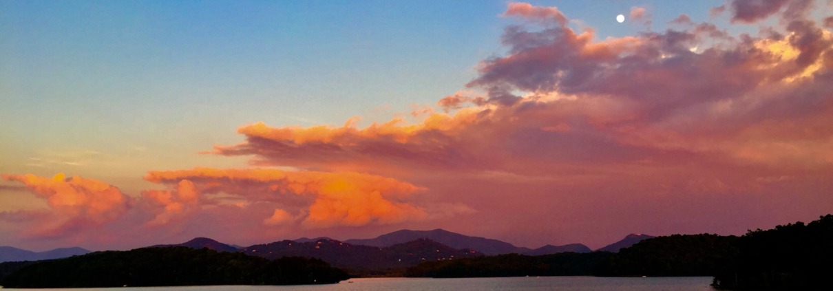 A panoramic sunset over Lake Chatuge with a full moon shining above vibrant pink and orange clouds.