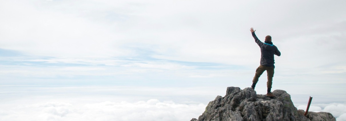 A man standing on a rocky mountain summit above a sea of clouds with one arm raised in surrender and worship, symbolizing emotional healing, trauma recovery, and spiritual liberation for a highly sensitive person.