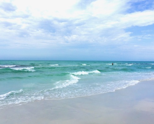 A peaceful beach scene on the Florida Gulf Coast featuring turquoise water with gentle waves lapping onto soft white sand under a bright sky with soft clouds, capturing the restoring essence of an HSP Breathwork Retreat with Benita Esposito.