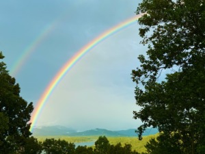 A vibrant double rainbow stretching across a calm lake and distant, misty blue mountains, framed naturally by green trees on the right and left borders.