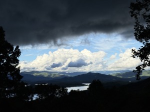 A dramatic storm cloud over a calm lake, with dark grey clouds on the upper left parting to reveal bright white clouds and a soft sunlight glow below.