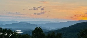 A panoramic sunrise view overlooking the rolling, misty ridges of the Blue Ridge Mountains with a calm lake visible in the valley below under a soft peach and orange sky.