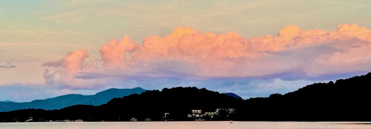 A wide panoramic sunset over the rolling, layered Blue Ridge Mountains, with dark tree silhouettes in the foreground and a soft peach and orange sky reflecting on a distant lake valley.